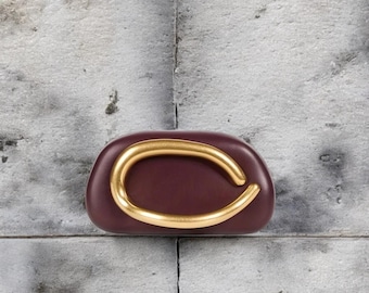 May include: A smooth, dark burgundy, pebble-shaped object featuring a prominent, curved, matte gold-toned metal accent piece centred on its front surface. The object has a rounded, organic form with a solid, uniform colour finish.