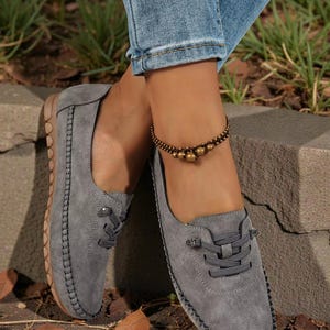 Puede incluir: Zapatos grises de ante sin cordones con cordones decorativos y un borde cosido. Los zapatos se combinan con vaqueros azules y una pulsera de tobillo con cuentas. Las suelas son de color marr&oacute;n claro.