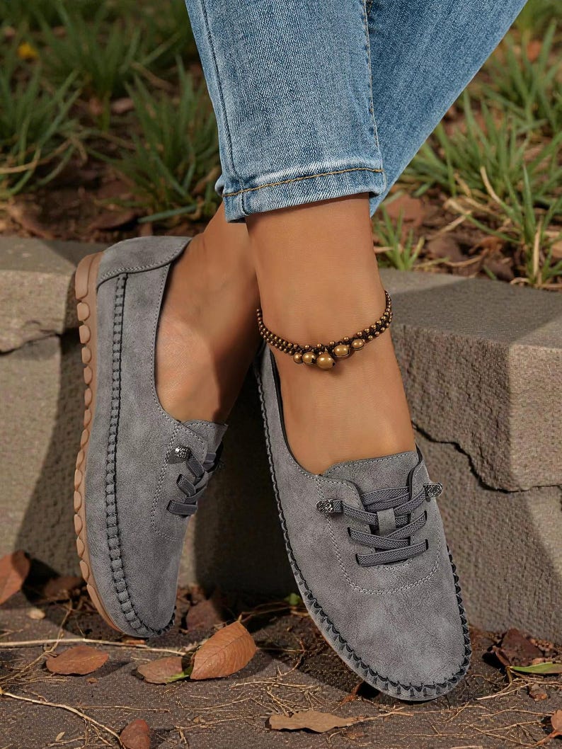 Puede incluir: Zapatos grises de ante sin cordones con cordones decorativos y un borde cosido. Los zapatos se combinan con vaqueros azules y una pulsera de tobillo con cuentas. Las suelas son de color marr&oacute;n claro.