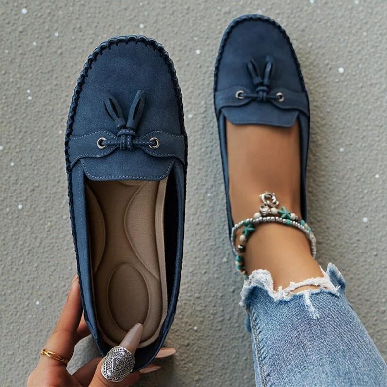 May include: A pair of blue suede loafers with decorative tassels and stitching. One shoe is shown from above, the other is being worn with a denim-clad leg and an anklet. The shoes have a rounded toe and a slip-on design.