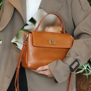 May include: A brown leather satchel handbag with a top flap closure and a gold metal clasp. The bag has a top handle and a long adjustable shoulder strap.