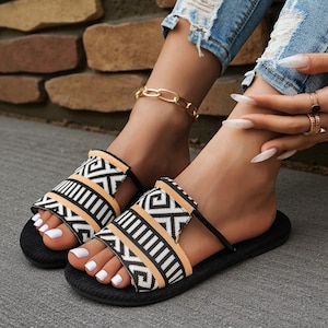 May include: Black and tan patterned slide sandals with a geometric design. The sandals have a black sole and straps. The sandals are worn by a person with white nail polish and a gold chain ankle bracelet.