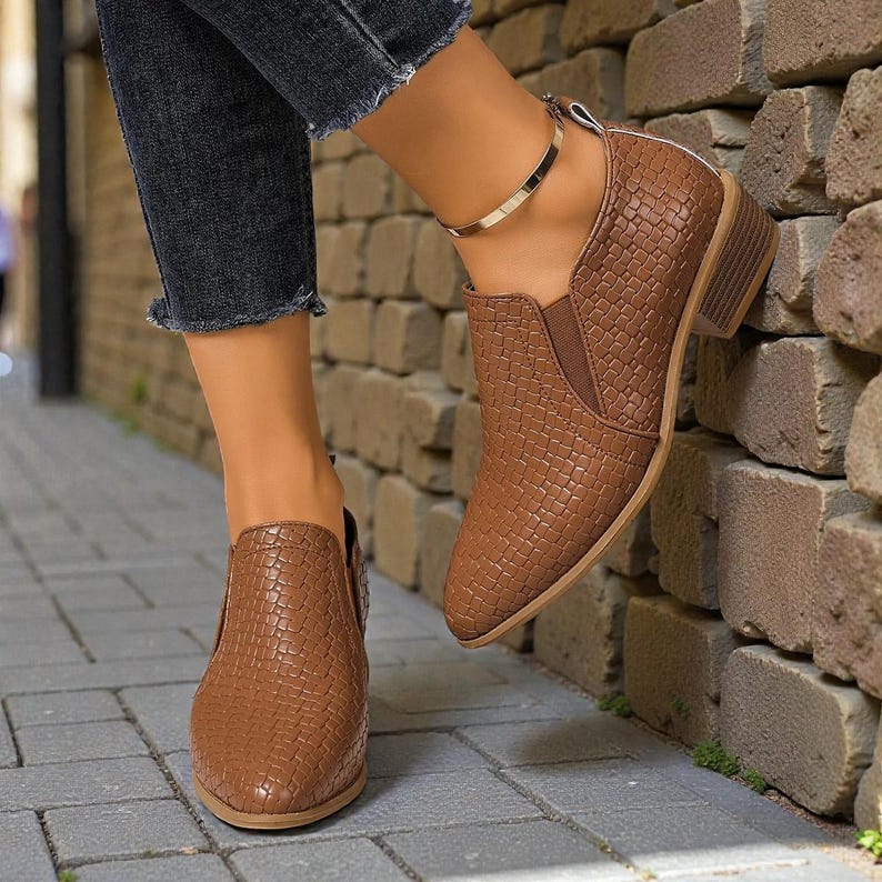 Pu&ograve; includere: Stivaletti marroni con una trama intrecciata. Le scarpe hanno un tacco basso e una punta a punta. Gli stivaletti sono indossati con jeans scuri e un braccialetto alla caviglia argentato. Le scarpe sono in materiale simile alla pelle.