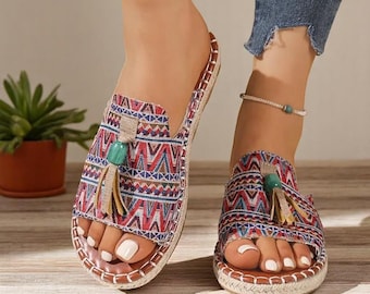 May include: Sandals with an open toe, featuring a vibrant geometric pattern in red, blue, and white. They have a jute sole and a small tassel with a turquoise bead. The design is repeated on the top of the sandal.