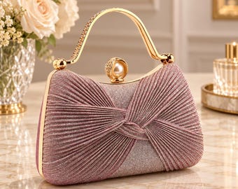 May include: A mauve-coloured clutch bag with a pleated, shimmering fabric texture. The bag features a gold-toned metal handle and a central clasp adorned with a single white pearl-like bead. The design includes a knotted fabric detail.