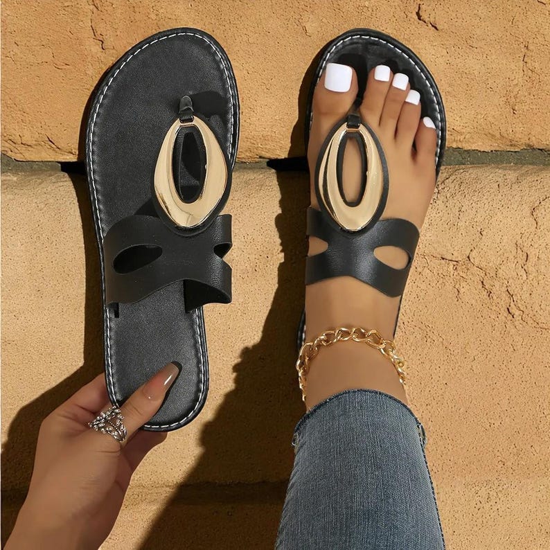 May include: Black sandals with a gold oval accent. The sandals have a thong-style design with a strap that wraps around the ankle. The sandals are shown on a brick surface.