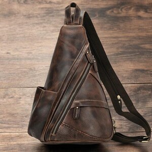 May include: A brown leather backpack with a triangular shape and multiple zippered compartments. The backpack has a single adjustable strap.