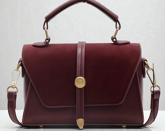 May include: A deep burgundy handbag with a smooth matte finish, featuring a top handle and a vertical strap closure secured by a circular gold-toned metal button. The bag includes gold-toned hardware, side rings for a shoulder strap, and a pointed metal tip.