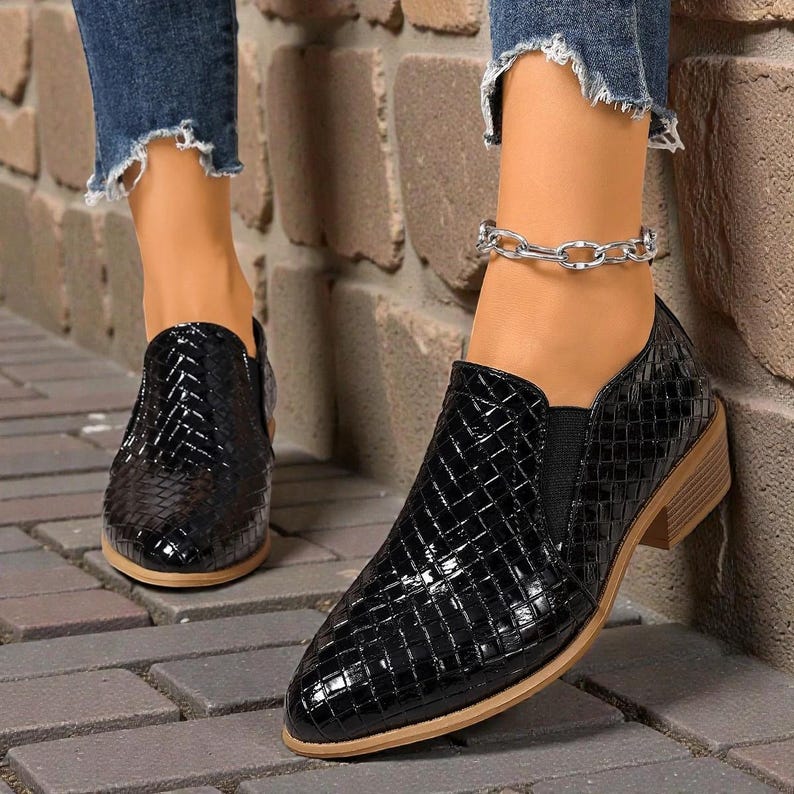 Pu&ograve; includere: Scarpe nere slip-on con un motivo intrecciato. Le scarpe hanno un tacco basso e una punta a punta. Le scarpe sono realizzate in materiale lucido. Le scarpe sono indossate con jeans blu.