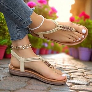 Könnte beinhalten: Beige Sandalen mit dekorativen Holzperlen und einem Zehensteg-Design. Die Sandalen haben eine braune Sohle und beige Riemen. Eine goldene Kette und ein weißes Perlenarmband sind ebenfalls sichtbar.