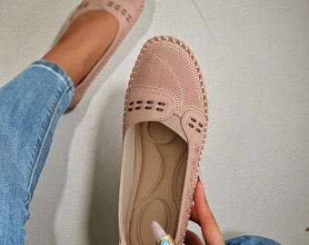 May include: Blush-coloured slip-on shoes with perforated detailing. The shoes have a rounded toe and a textured design on the upper. The sole is a lighter shade of beige. The shoes are shown with blue jeans.