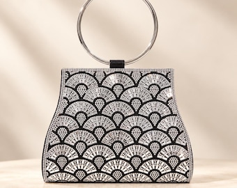 May include: A black handbag featuring a repeating silver fan-shaped pattern created with small crystals and rectangular rhinestones. The bag has a structured trapezoidal shape and a single circular silver metal handle attached at the top centre.