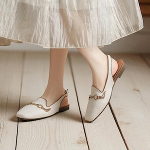 May include: Off-white slingback loafers with a square toe and gold-tone hardware. The shoes have a woven texture and brown leather accents. The soles are brown, and the shoes have a low heel.