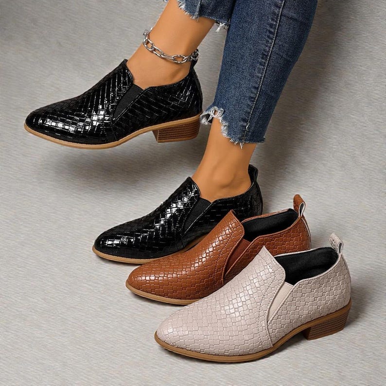 Pu&ograve; includere: Scarpe slip-on nere, marroni e beige con una trama intrecciata. Le scarpe hanno un tacco basso e una punta a punta. La scarpa nera &egrave; indossata. Le scarpe sono esposte su una superficie grigio chiaro.