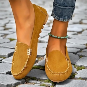 以下が含まれることがあります： マスタードイエローのスエードローファー、ゴールドカラーのハードウェアアクセント付き。丸いつま先と装飾的なステッチが特徴です。画像は石の表面に置かれた靴を示しています。
