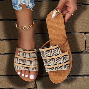 May include: A pair of brown corduroy sandals with woven fabric straps featuring a geometric pattern in beige, black, and orange. One sandal is being held up, while the other is on a foot with a gold chain anklet.