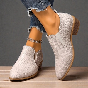 Pu&ograve; includere: Stivaletti grigio chiaro con una trama intrecciata. Le scarpe con tacco basso hanno la punta arrotondata e un design slip-on. Le suole sono di colore marrone chiaro. Gli stivali sono mostrati con jeans blu.
