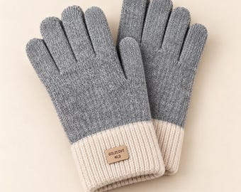 May include: A pair of grey knit gloves featuring a ribbed cream-coloured cuff. A small rectangular tan patch is stitched onto the cuff of the left glove, displaying the black text GOLDLOVE NLD in a clear, sans-serif font.