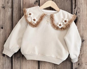 May include: A cream-coloured long-sleeve sweatshirt featuring a large, rounded collar with brown ruffled trim and white crocheted flower appliqués. The garment includes a short front placket with two buttons and ribbed cuffs at the sleeves and hem.