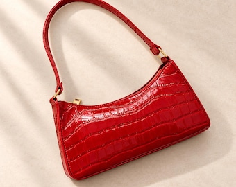 May include: A bright red shoulder bag featuring a glossy, embossed crocodile skin texture. The handbag has a slim, rectangular silhouette, a single fixed strap, and a gold-toned metal zip closure along the top edge for secure storage.