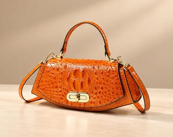 May include: A vibrant orange handbag with a textured crocodile-embossed pattern. The bag features a curved silhouette, a single top handle, a detachable shoulder strap, and a polished gold-tone metal turn-lock closure on the front.