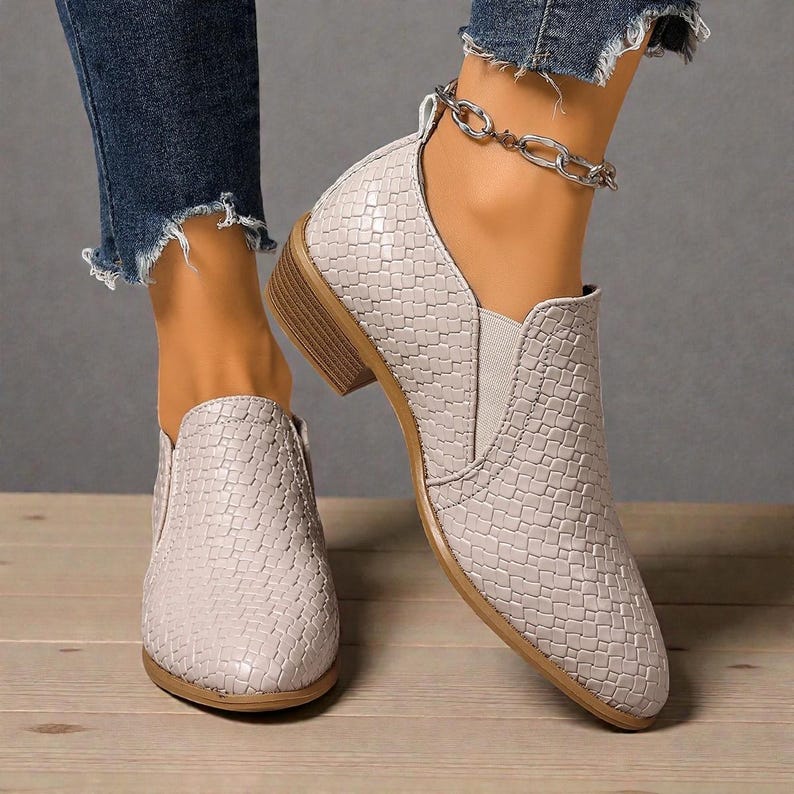 Pu&ograve; includere: Un paio di stivaletti grigio chiaro con una trama intrecciata. Le scarpe hanno un tacco basso e un design slip-on. Le suole sono marrone chiaro. Gli stivaletti sono indossati con jeans blu.
