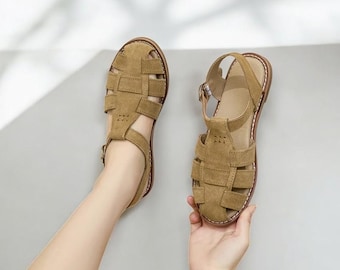 May include: Two tan suede sandals with a woven design. The sandals have a strap around the ankle and a buckle closure. The soles are brown with white stitching. The sandals are displayed on a light gray background.