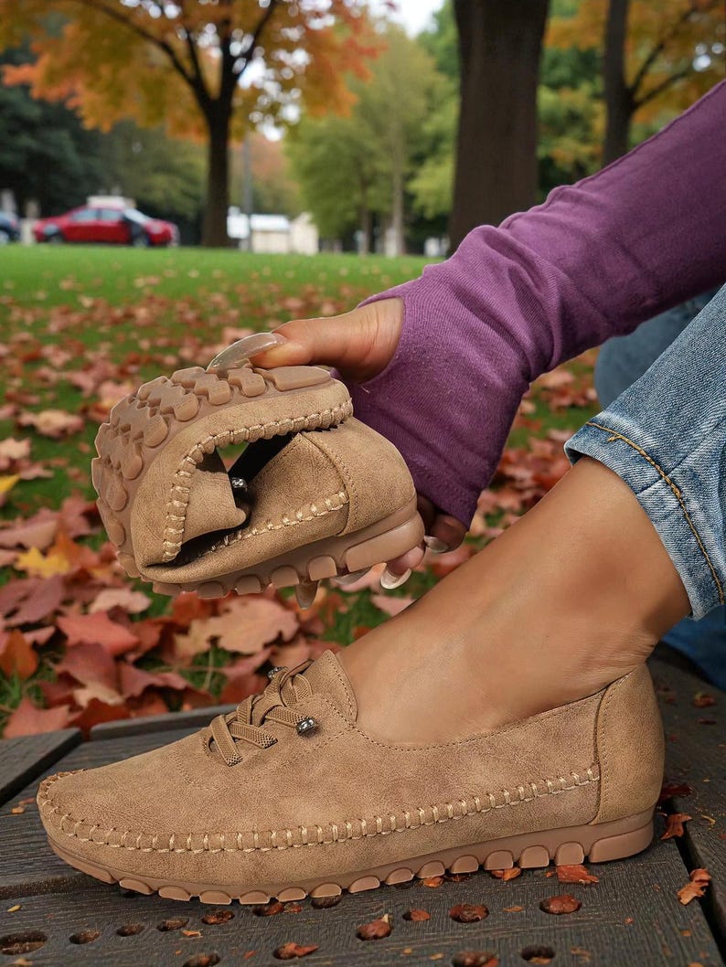 Puede incluir: Mocasines color beige con aspecto de ante, con costuras y suelas de goma. Los zapatos tienen cordones y un peque&ntilde;o elemento met&aacute;lico decorativo. Uno de los zapatos est&aacute; doblado, mostrando su flexibilidad. El fondo muestra hojas de oto&ntilde;o.
