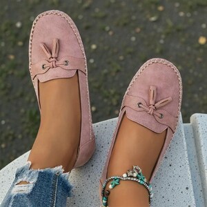 May include: A pair of dusty rose-colored loafers with tassel details. The shoes have a rounded toe and a stitched edge. The loafers are made of a soft material and are designed for comfort and style. The shoes are a slip-on style.