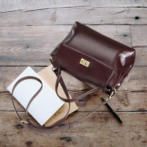 May include: A brown leather satchel bag with a gold clasp and a long adjustable strap. The bag is lying on a wooden surface with a notebook and pen nearby.