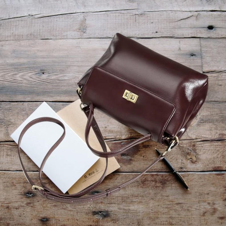 May include: A brown leather satchel bag with a gold clasp and a long adjustable strap. The bag is lying on a wooden surface with a notebook and pen nearby.