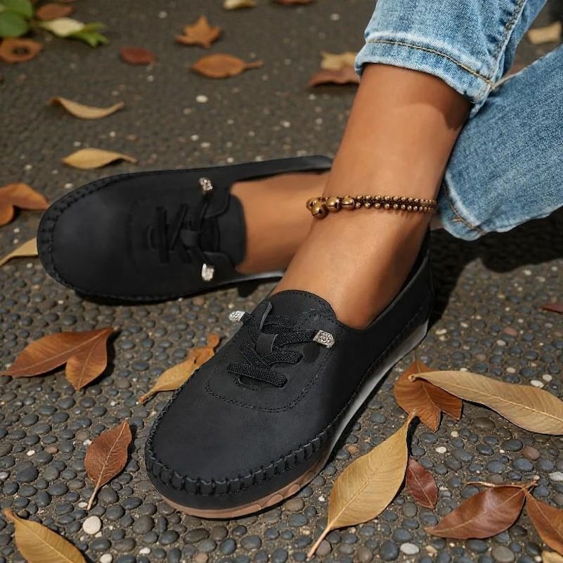 Puede incluir: Zapatos negros casuales con dise&ntilde;o de cordones y costuras decorativas alrededor de la suela. Los zapatos tienen una punta redondeada y un perfil bajo. Tambi&eacute;n se ve una pulsera de tobillo dorada y hojas ca&iacute;das.