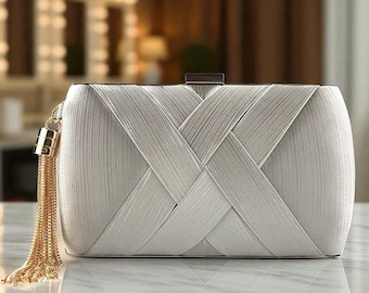 May include: A rectangular white clutch bag featuring a textured surface with a crisscross pattern. The bag includes a gold-toned metal clasp at the top and a long, dangling gold-toned chain tassel attached to the side of the frame.