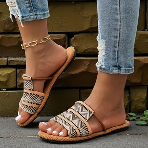 May include: A pair of open-toe sandals with a geometric pattern in black, beige, and orange. The sandals have a brown strap and a tan sole. The person is wearing light blue denim jeans with distressed details and a gold chain anklet.