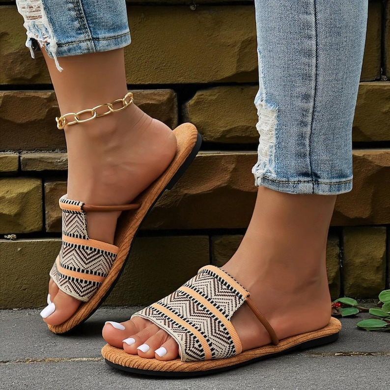 May include: A pair of open-toe sandals with a geometric pattern in black, beige, and orange. The sandals have a brown strap and a tan sole. The person is wearing light blue denim jeans with distressed details and a gold chain anklet.