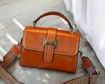 May include: A structured cognac-coloured leather handbag featuring a top handle, a rectangular flap with a central gold-tone metal clasp, and a wide, detachable woven shoulder strap with a multi-coloured chevron pattern and white stitching.