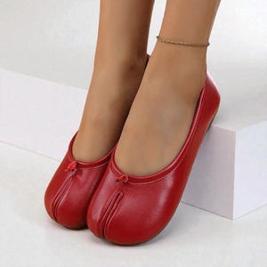 May include: Red ballet flats with a split-toe design. The shoes have a small bow detail at the top and a rounded toe. The flats are made of a shiny material and are shown on a white surface.