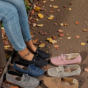 Puede incluir: Se muestran varios zapatos sin cordones de ante en negro, azul marino, gris, rosa y marr&oacute;n. Los zapatos informales tienen cordones y una puntera redonda. La imagen tambi&eacute;n muestra a una persona que lleva vaqueros azules.