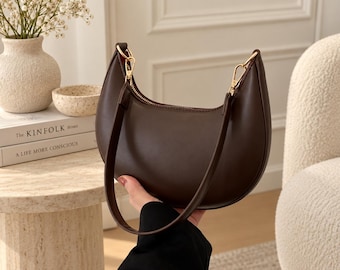 May include: A dark brown crescent-shaped shoulder bag with a smooth finish, featuring a gold-toned zip closure along the top and a matching brown strap attached with gold-toned metal clasps on both sides of the bag.