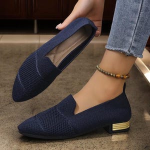 Può includere: Scarpe slip-on blu navy con tomaia in maglia a rete e un piccolo tacco a blocco con un accento dorato. Una scarpa è tenuta in alto, mentre l'altra è indossata su un piede. Le scarpe hanno la punta a punta e un collo del piede basso.