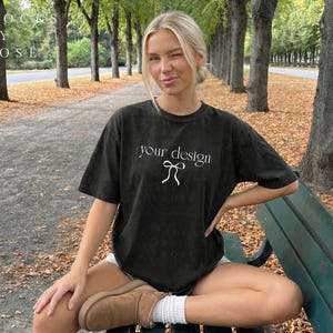 May include: A black t-shirt with the text "your design" in white, featuring a bow graphic. The model is sitting on a park bench, wearing white shorts and brown boots. The background includes trees and fallen leaves.