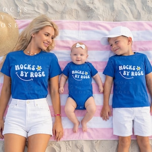 May include: A family of three wearing matching blue t-shirts and a onesie with the text "MOCKS BY ROSE" on a pink and white striped beach towel. The mother wears white shorts, and the son wears white shorts and a white cap. A straw hat is in the background.
