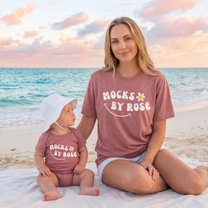 May include: A woman and a baby wearing matching mauve t-shirts and sitting on a beach. The shirts read "MOCKS BY ROSE" in white text with a daisy and a smiling face. The baby wears a white sun hat. The background shows the ocean and a colorful sky.