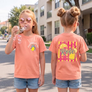 May include: Two young girls wearing peach-colored t-shirts and denim shorts stand on a street. One blows bubbles, while the other's shirt features a graphic with the text "Rose" and "She Be Makin' Mockups."