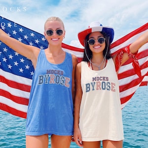 Può includere: Due persone con occhiali da sole e canottiere con la scritta "MOCKS BY ROSE" davanti a una bandiera americana. Una canotta è blu, l'altra color crema. La persona a destra indossa un cappello rosso, bianco e blu.