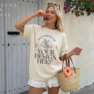 May include: Cream-colored t-shirt with the text "YOUR DESIGN HERE" and a circular logo. The model is holding a straw tote bag with a pink flower and cherries. The shirt is paired with a white skirt.