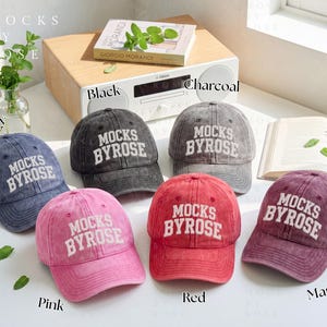 Könnte beinhalten: Sechs Baseballkappen in den Farben Marineblau, Schwarz, Anthrazit, Pink, Rot und Weinrot. Jede Kappe trägt den weißen Schriftzug "MOCKS BYROSE". Die Kappen sind auf einer weißen Oberfläche angeordnet. Ein Buch und eine Pflanze sind ebenfalls zu sehen.