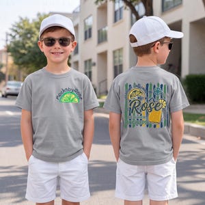 May include: A young person wearing a gray t-shirt and white shorts. The front of the shirt has a lime graphic and the text "mocksbyrose". The back of the shirt has a graphic with a lemon, a beer mug, and the text "Rose Mockups".