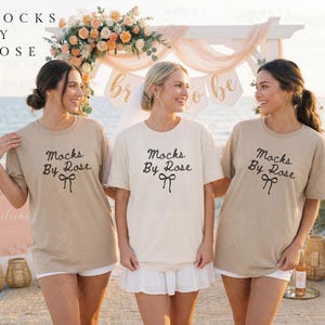 Könnte beinhalten: Drei Frauen tragen T-Shirts mit dem Text "Mocks By Rose" in schwarzer Schrift. Die Shirts sind in Beige- und Cremetönen gehalten. Die Frauen stehen an einem Strand mit einem Blumengewölbe im Hintergrund.