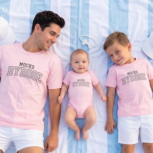 May include: A family of three wearing matching pink t-shirts and a onesie with the text "MOCKS BYROSE" in a collegiate font. The man and boy are wearing white shorts, and the baby is wearing a pink onesie. White hats are placed on the striped background.